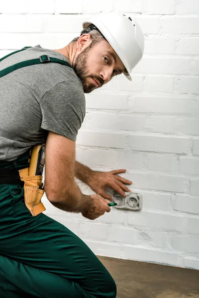 Handsome electrician repairing and unscrewing power socket with screwdriver at home, looking at camera — Stock Photo