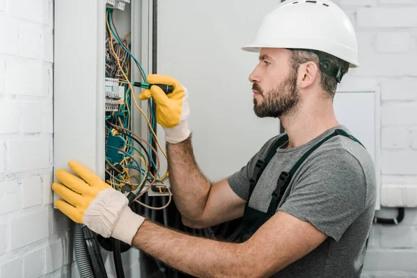 Side view of handsome bearded electrician repairing electrical box and using screwdriver in corridor — Stock Photo