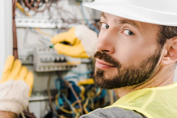 Beau barbu électricien réparer la boîte électrique, en utilisant un tournevis dans le couloir et en regardant la caméra — Photo de stock