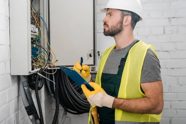 Side view of serious electrician holding clipboard and checking electrical box in corridor — Stock Photo