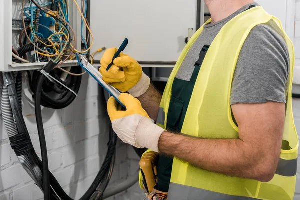 Abgeschnittenes Bild eines Elektrikers, der Klemmbrett und Stift in der Nähe eines Stromkastens im Flur hält — Stockfoto