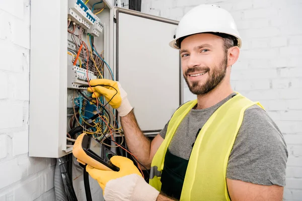 Smiling handsome electrician checking electrical box with multimetr in corridor and looking at camera — Stock Photo