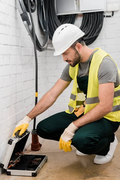 Handsome electrician squatting near electrical box and looking into toolbox in corridor — Stock Photo