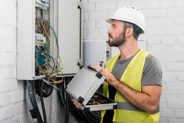 Side view of handsome electrician holding toolbox and looking at electrical box in corridor — Stock Photo