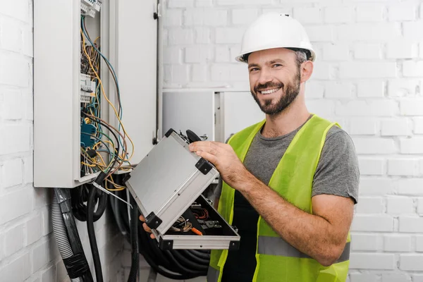 Electricista sonriente sosteniendo la caja de herramientas cerca de la caja eléctrica en el pasillo y mirando a la cámara - foto de stock