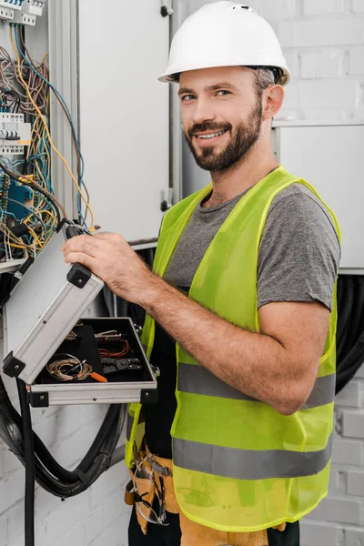 Sonriente electricista guapo sosteniendo caja de herramientas cerca de la caja eléctrica en el pasillo y mirando a la cámara - foto de stock