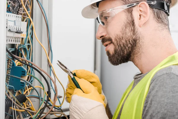Side view of handsome electrician holding pliers and looking at electrical box in corridor — Stock Photo