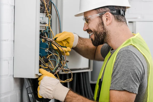Side view of smiling handsome electrician repairing electrical box with pliers in corridor — Stock Photo