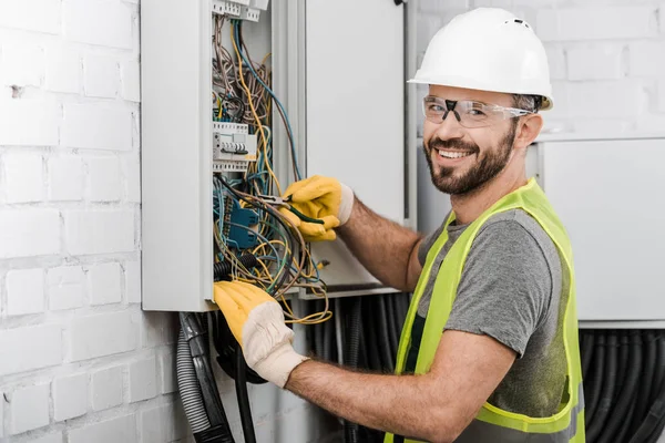 Smiling handsome electrician repairing electrical box with pliers in corridor and looking at camera — Stock Photo
