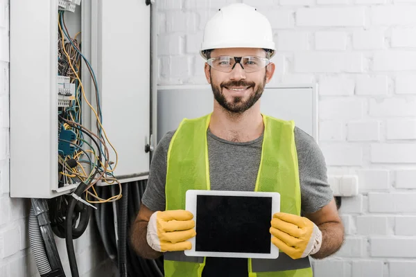 Eletricista bonito sorrindo mostrando tablet com tela em branco perto de caixa elétrica no corredor — Fotografia de Stock