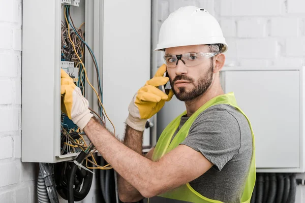 Electricista sorprendido comprobando la caja eléctrica y hablando por teléfono inteligente en el pasillo - foto de stock