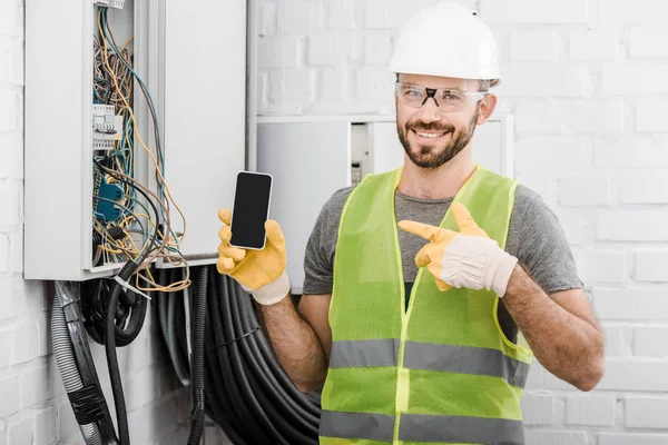 Electricista guapo sonriente apuntando en el teléfono inteligente con pantalla en blanco cerca de la caja eléctrica en el pasillo - foto de stock