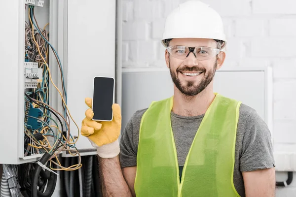 Electricista guapo sonriente mostrando teléfono inteligente con pantalla en blanco cerca de la caja eléctrica en el pasillo - foto de stock