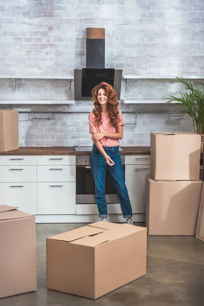 Attractive smiling woman with curly hair standing near cardboard boxes at new kitchen — Stock Photo
