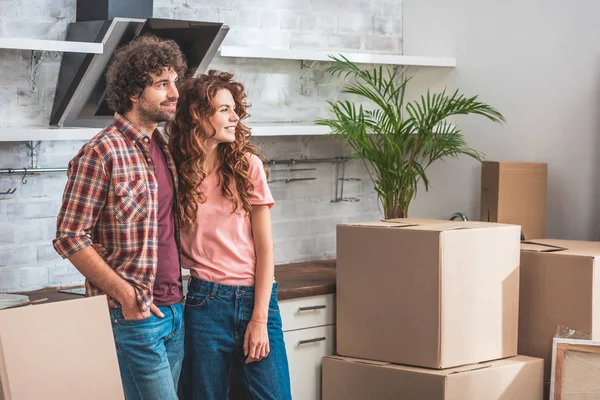 Пара с кудрявыми волосами, стоящими рядом с картонными коробками и смотрящими на новую кухню — стоковое фото