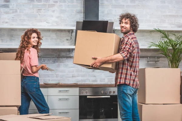 Feliz pareja desembalaje cajas de cartón juntos en la nueva cocina y mirando a la cámara - foto de stock