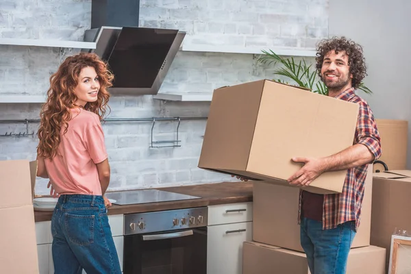 Sonriente pareja desembalaje cajas de cartón juntos en la nueva cocina y mirando a la cámara - foto de stock