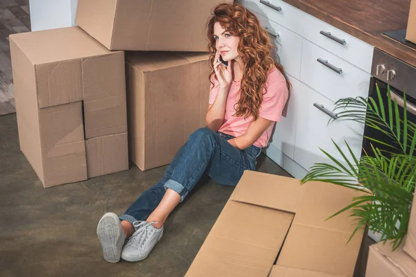 Vista de ángulo alto de la mujer atractiva con el pelo rizado sentado en el suelo cerca de cajas de cartón y hablando por teléfono inteligente en el nuevo hogar - foto de stock