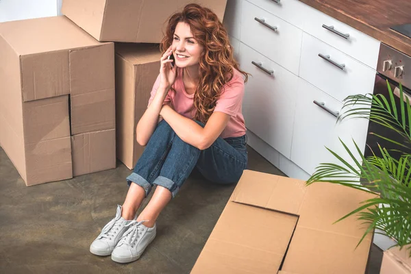 Smiling attractive woman with curly hair sitting on floor near cardboard boxes and talking by smartphone at new home — Stock Photo