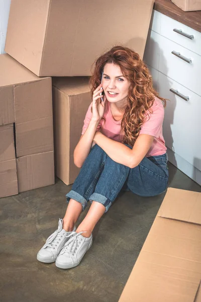 Glückliche schöne Frau mit lockigem Haar sitzt auf dem Boden neben Pappkartons und spricht im neuen Zuhause per Smartphone — Stockfoto