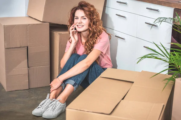 Smiling attractive woman sitting on floor near cardboard boxes and talking by smartphone at new home — Stock Photo