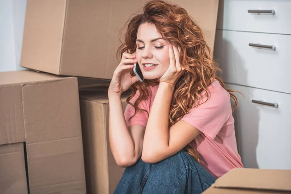 Cheerful attractive woman with curly hair sitting on floor near cardboard boxes and talking by smartphone at new home — Stock Photo