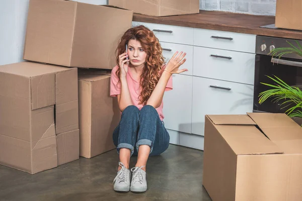 Mujer irritada con el pelo rizado sentado en el suelo cerca de cajas de cartón, hablando por teléfono inteligente y haciendo gestos en la nueva cocina - foto de stock