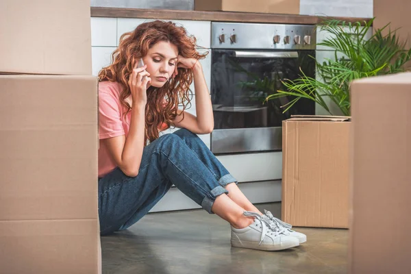 Donna triste con i capelli ricci seduta sul pavimento vicino a scatole di cartone e parlando da smartphone nella nuova cucina — Foto stock