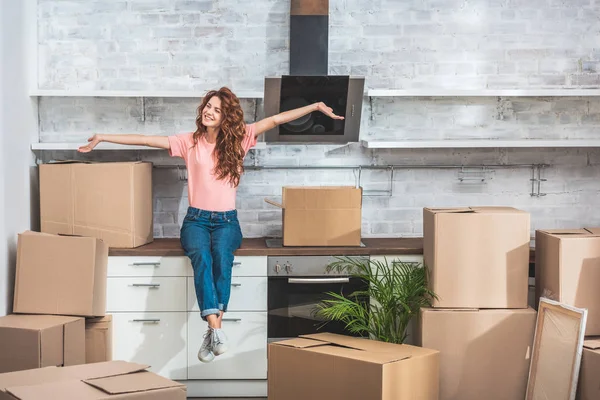 Mujer atractiva sonriente con el pelo rizado sentado en el mostrador de la cocina entre cajas de cartón con las manos extendidas en el nuevo hogar - foto de stock