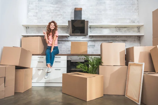 Mujer atractiva sonriente sentada en el mostrador de la cocina entre cajas de cartón y mostrando signo de paz en un nuevo hogar - foto de stock