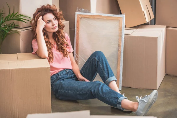 Attractive woman with curly red hair in casual clothes sitting on floor near cardboard boxes at new home — Stock Photo