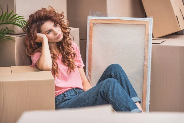 Tired beautiful woman with curly red hair sitting on floor near cardboard boxes at new home — Stock Photo
