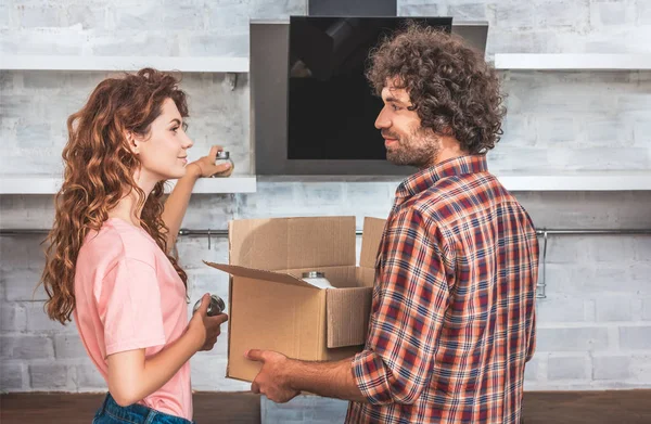 Vista lateral de la pareja tomando utensilio de la caja de cartón en el nuevo hogar y mirándose entre sí - foto de stock