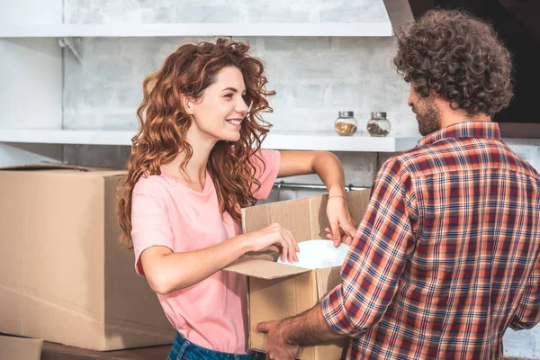 Улыбающаяся пара берет посуду из картонной коробки в новом доме и смотрит друг на друга — стоковое фото