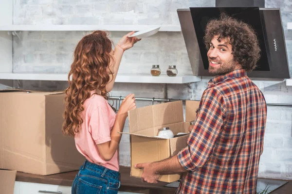 Sorrindo namorado segurando caixa de papelão, namorada colocando prato em prateleiras em nova casa — Fotografia de Stock