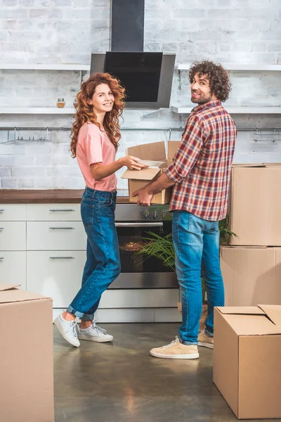 Smiling couple unpacking cardboard box together at new home and looking at camera — Stock Photo