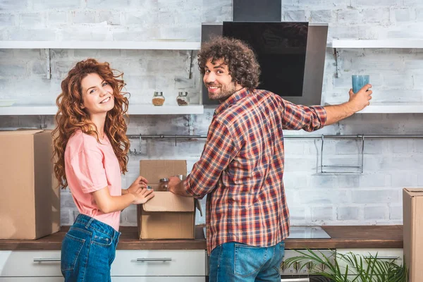 Sonriente pareja desembalaje cajas de cartón en el nuevo hogar y mirando a la cámara - foto de stock