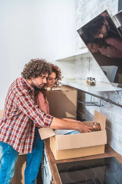 Vista lateral de la caja de cartón de abrazo y desembalaje de pareja en la cocina nueva - foto de stock