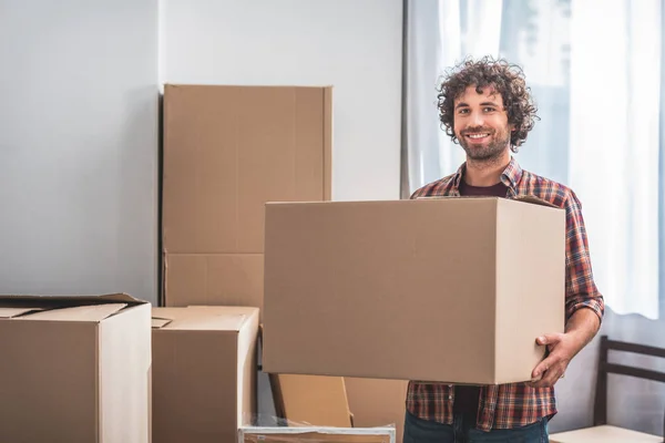 Sorrindo homem bonito com cabelo encaracolado segurando caixa de papelão em nova casa — Fotografia de Stock