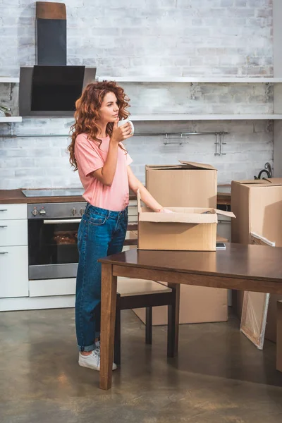 Hermosa mujer bebiendo café y desempacando caja de cartón en el nuevo hogar - foto de stock