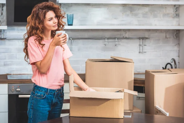 Attractive woman holding cup of tea and unpacking cardboard box at new home — Stock Photo