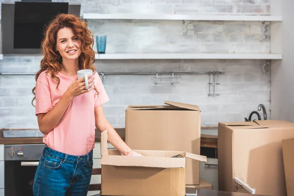 Bella donna sorridente che tiene la tazza di caffè e disimballaggio scatola di cartone a nuova casa — Foto stock