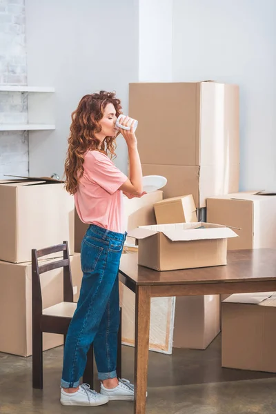 Bela mulher beber café e desembalagem caixa de papelão em nova casa — Fotografia de Stock