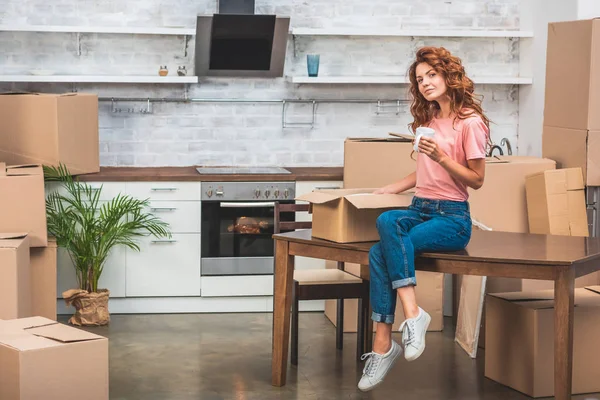 Schöne Frau mit Tasse Kaffee und Pappschachtel auf dem Tisch im neuen Zuhause auspacken — Stockfoto