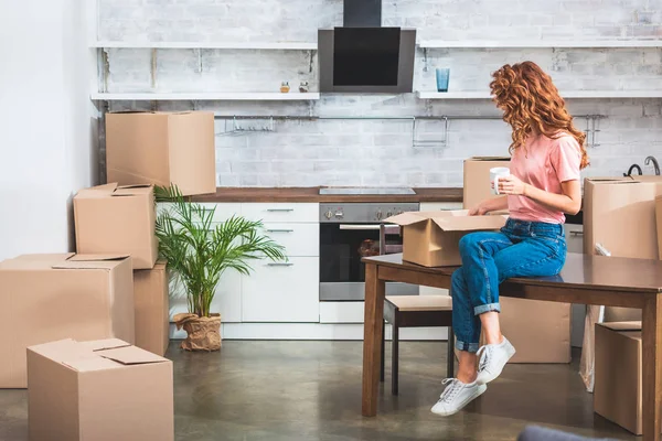 Side view of woman holding cup of coffee and unpacking cardboard box on table at new home — Stock Photo