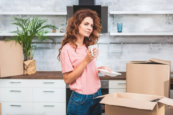 Beautiful woman with curly red hair holding cup of coffee and plate at new home — Stock Photo