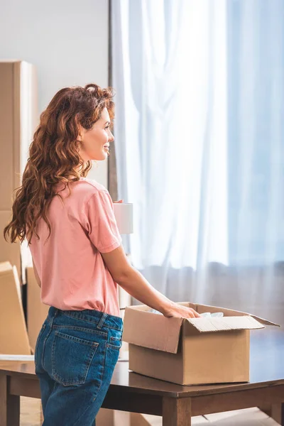 Vista lateral da mulher bonita com cabelo encaracolado vermelho segurando xícara de café e desembalagem caixa de papelão na nova casa — Fotografia de Stock