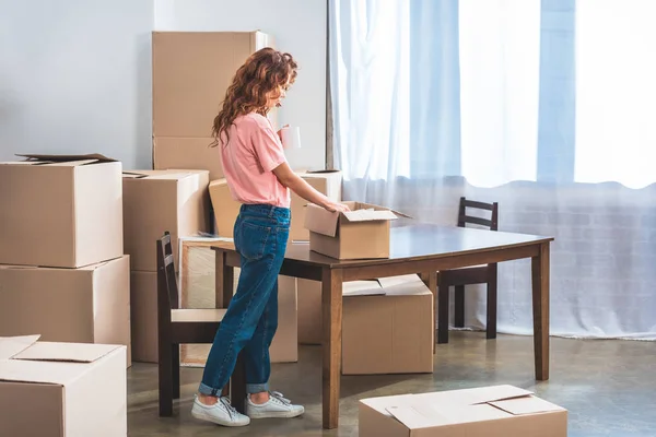 Vista lateral de la hermosa mujer con pelo rojo rizado desembalaje cajas de cartón en el nuevo hogar - foto de stock