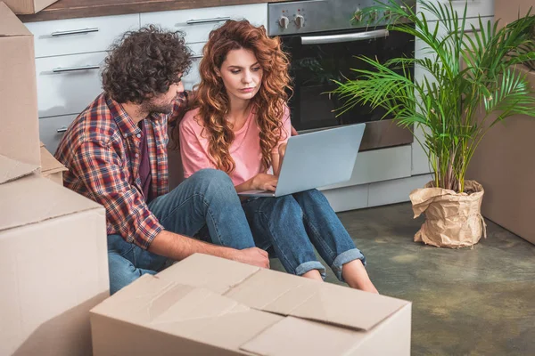 Pareja con el pelo rizado sentado con el ordenador portátil cerca de cajas de cartón en el suelo en la cocina nueva - foto de stock