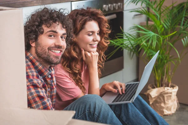 Smiling couple sitting on floor with laptop near cardboard boxes in new kitchen — Stock Photo
